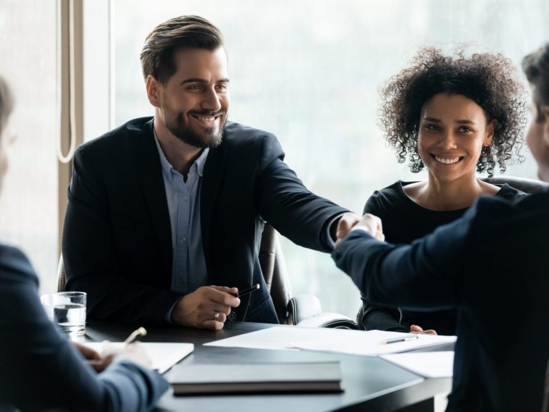 Happy successful businessman in suit shaking hand of business partner at company meeting. Diverse team fasten deal with gesture. Leader in suit with african american assistant congratulates clients.